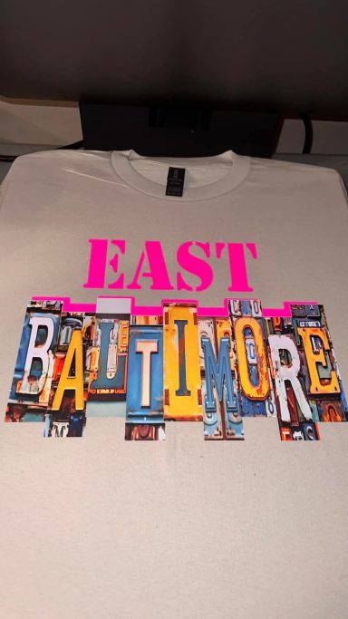 A light gray t-shirt with bold, colorful "EAST BALTIMORE" graphic design.